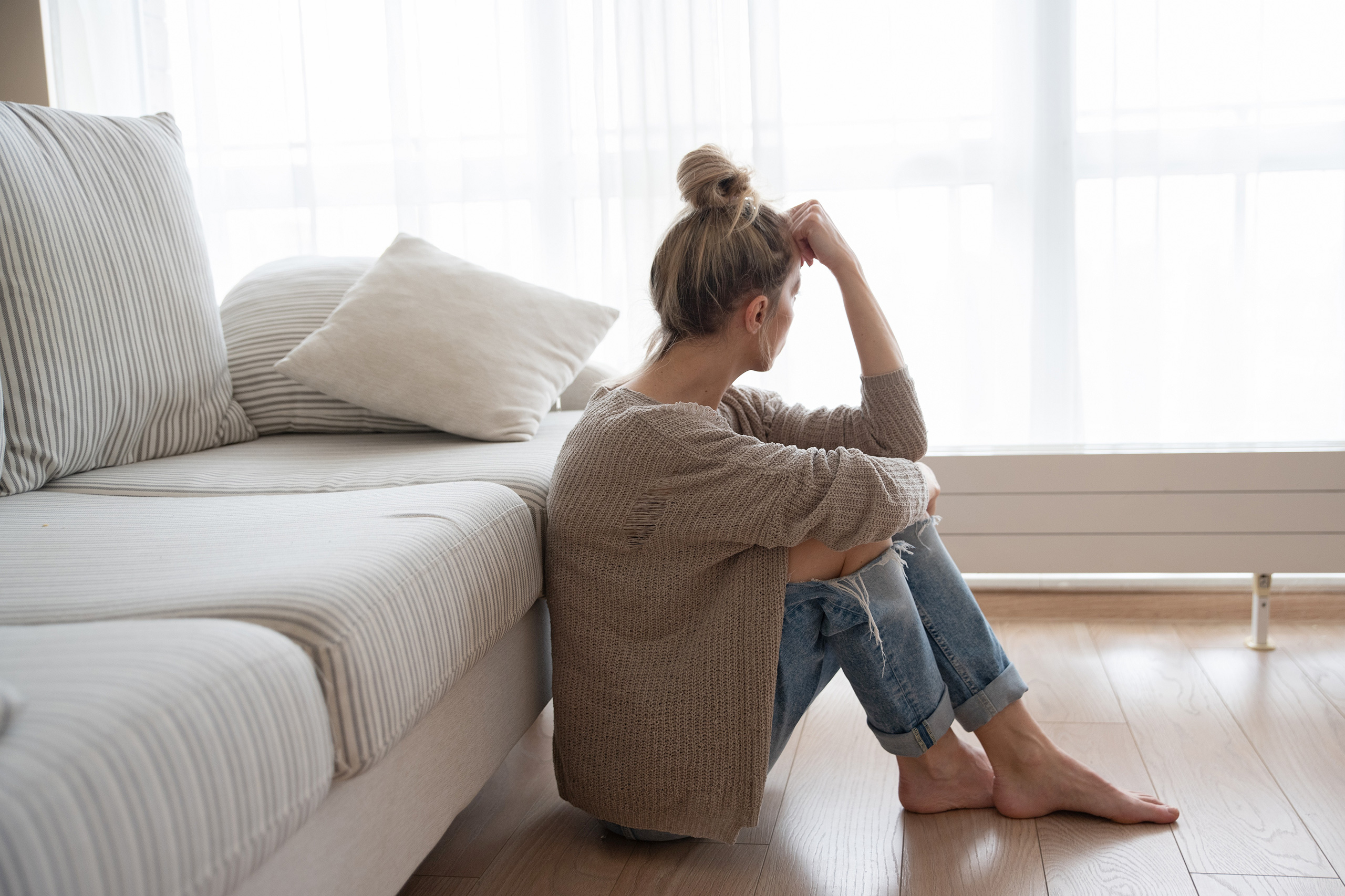 Eine Frau sitzt am Boden vor der Couch in einem Wohnzimmer, die Arme auf die angezogenen Knie gestützt und schaut gedankenverloren Richtung Fenster