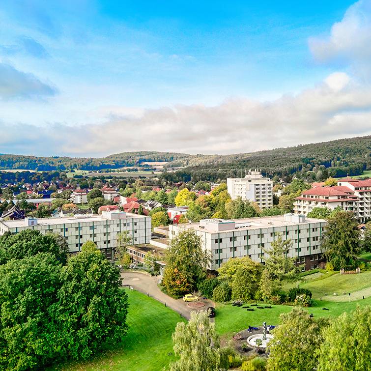 Luftaufnahme einer Reha-Klinik inmitten grüner Landschaft mit umliegenden Hügeln, Feldern und einem kleinen Ort im Hintergrund.