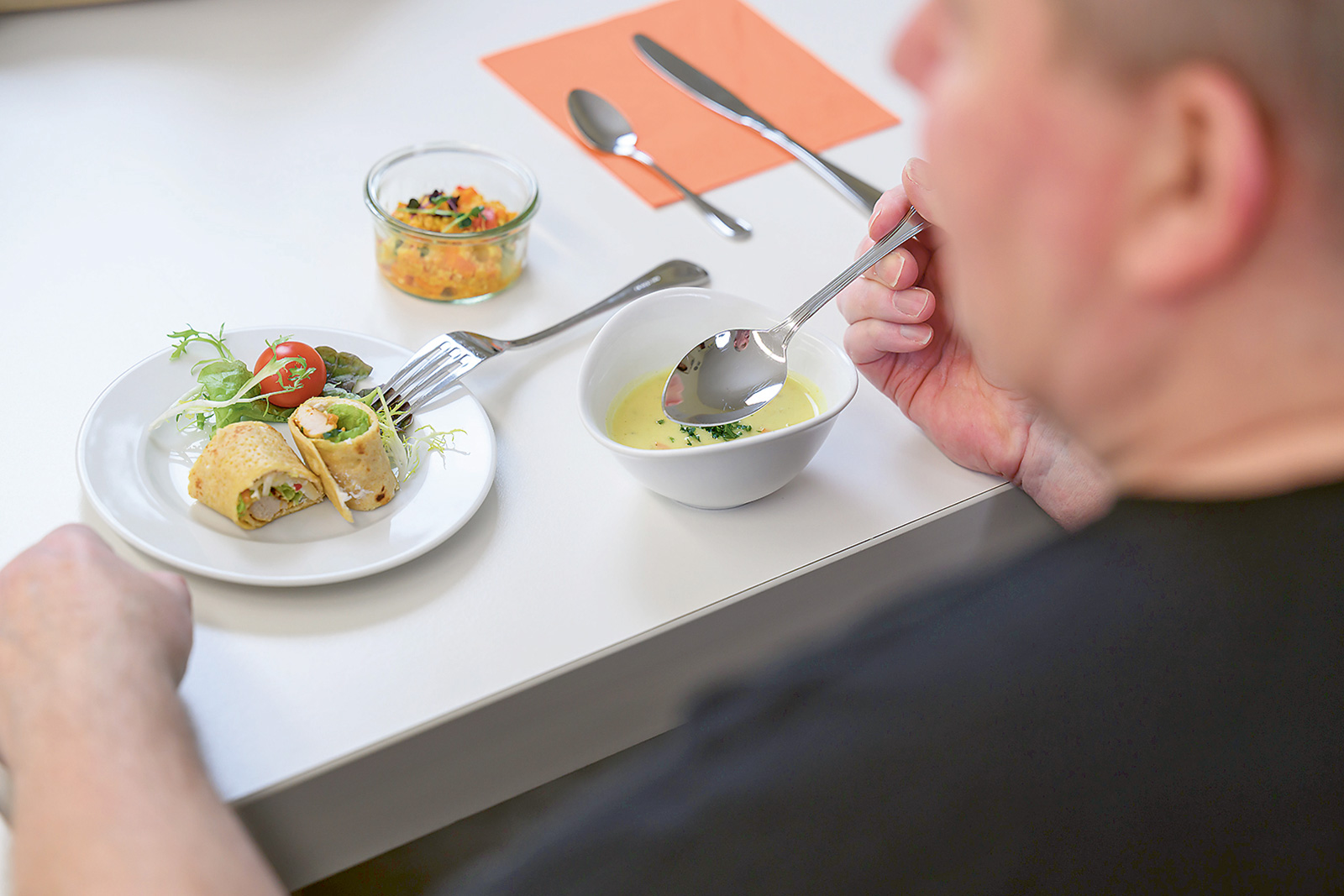 Mann isst Suppe und Wraps mit Salat an einem gedeckten Tisch, daneben ein Glas mit Gemüse – gesunde Mahlzeit in heller Umgebung.