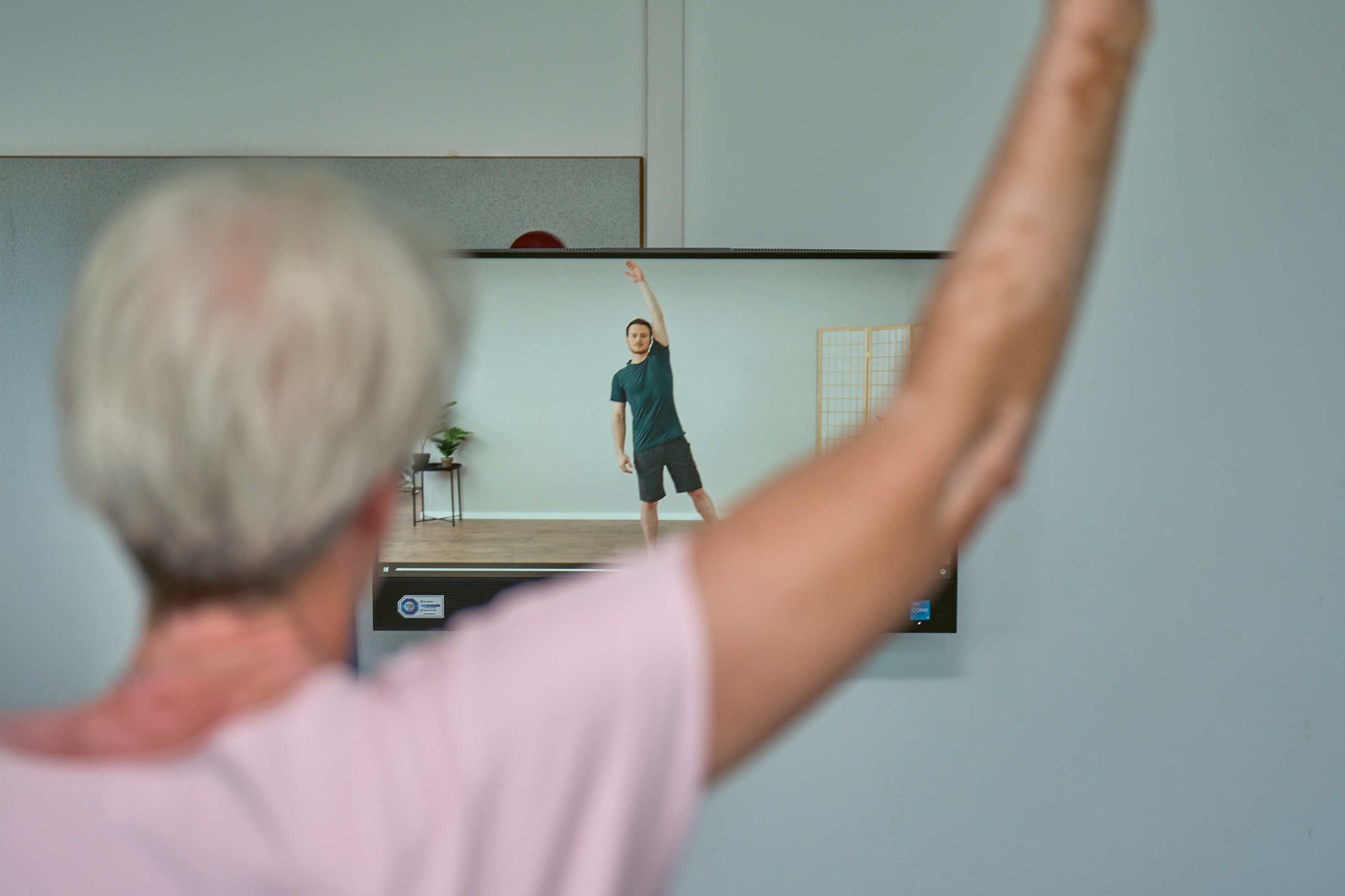 Ein älterer Mensch macht Gymnastikübungen nach, vorne im Bild unscharf, im Hintergrund sieht man einen Therapeuten, der Bewegungen vormacht.