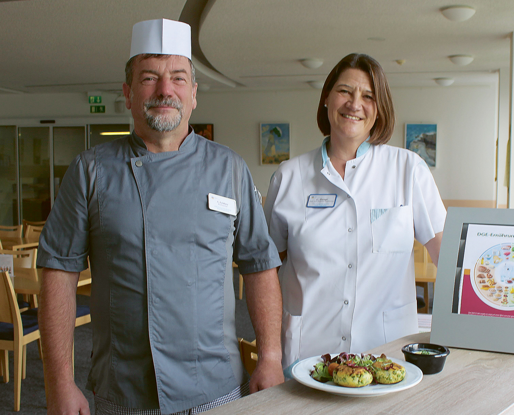 Zwei Mitarbeitende aus der Reha-Klinikküche, ein Mann und eine Frau mit Kochkleidung, präsentieren gemeinsam ein Gericht – Fokus auf gesunde Ernährung und Nachhaltigkeit.